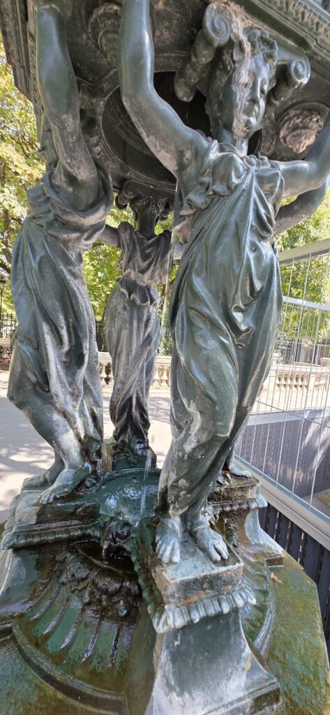 Dark green cast iron Wallace fountain in Paris, four draped female figures with raised arms holding up the dome, water falling into the basin at their feet