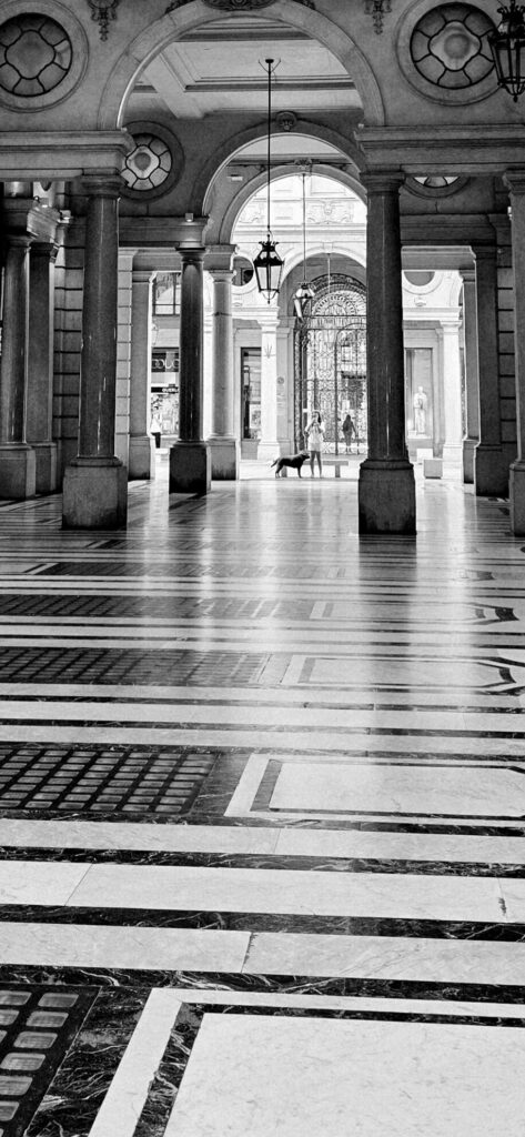 Black and white view along a grand arcade with stone columns and arches, hanging lanterns, an inlaid marble floor in bold geometric bands running away from the camera, and a small dog and two distant figures at the far end
