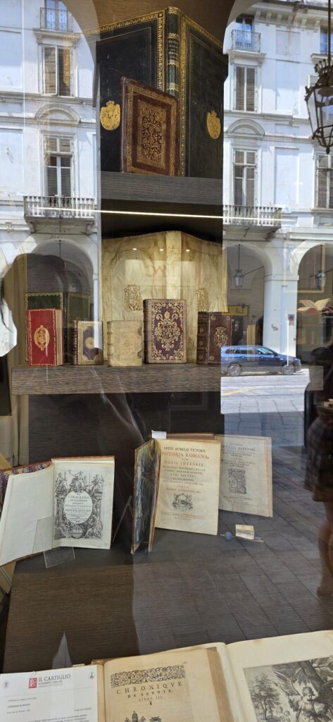 An antiquarian bookshop window with old leather and vellum bound volumes standing on shelves and two large books lying open showing engraved title pages, with the arcade and a pale palace facade reflected across the glass