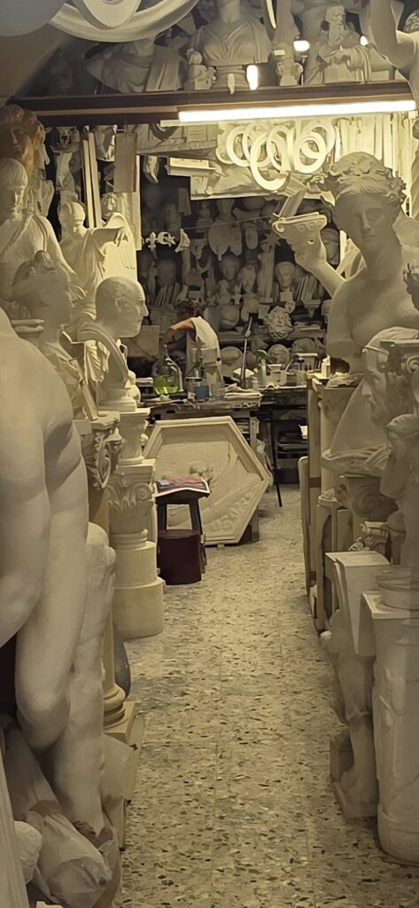 The inside of a plaster cast workshop packed floor to ceiling with white casts of torsos, heads, hands, columns and architectural mouldings, narrowing to a workbench at the back where somebody is bent over working