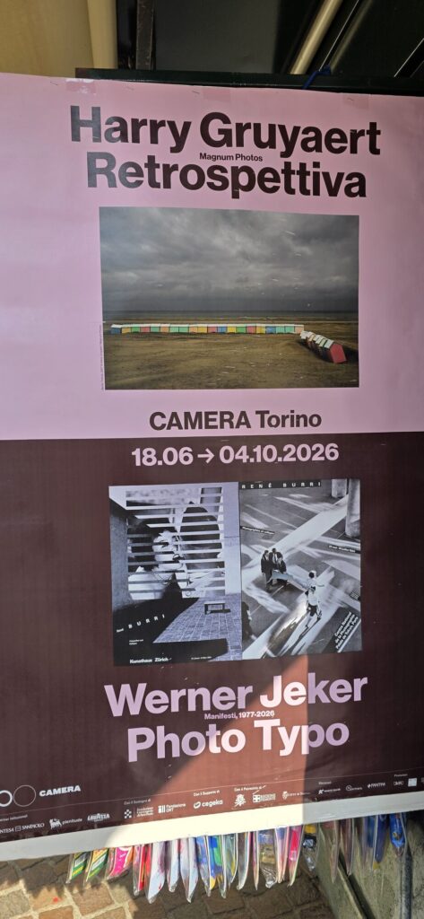 A tall printed banner hanging under an arcade advertising Harry Gruyaert Retrospettiva at CAMERA Torino with dates, and below it a second panel for Werner Jeker Photo Typo