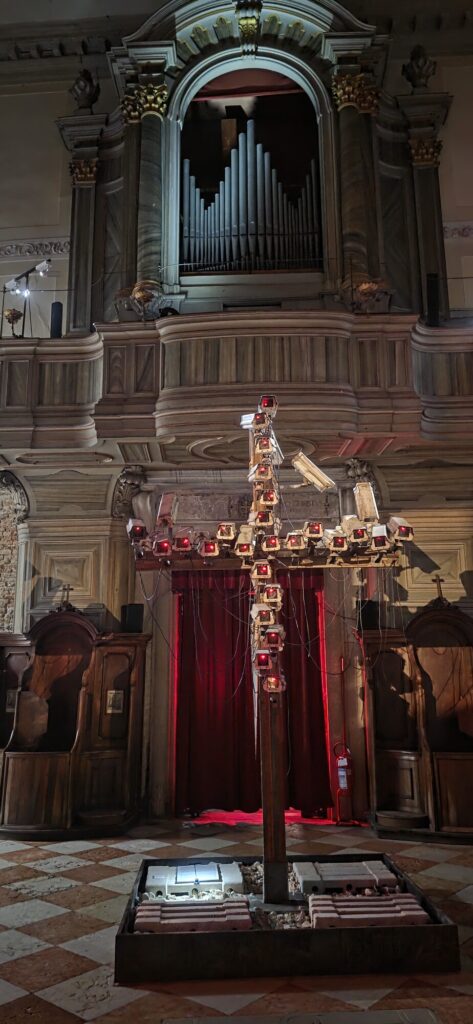 A tall wooden cross standing in a dim church, its arms and upright covered in dozens of security cameras with glowing red lights, a church organ and red curtain behind it and a low bed of rubble at its foot