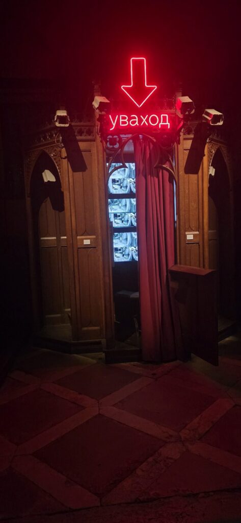 A dark wooden confessional in a dim church with a red velvet curtain across the middle bay, red neon above it reading a Belarusian word with a downward arrow, security cameras mounted on the woodwork and monitor screens glowing inside the grille