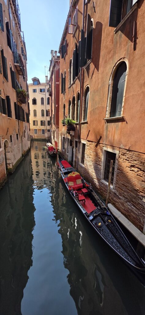 A narrow Venetian canal between ochre and terracotta walls with a moored gondola and its red upholstered seats on the right, the buildings reflected almost perfectly in still green water