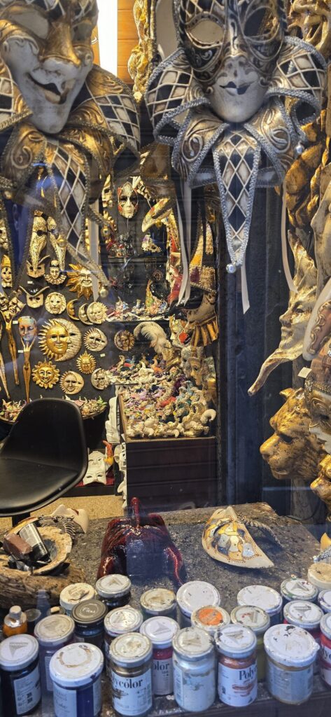 Through a shop window, a mask maker's workbench crowded with dozens of open pots of acrylic paint and brushes, with gilded jester masks, sun faces and animal masks hanging on the wall behind