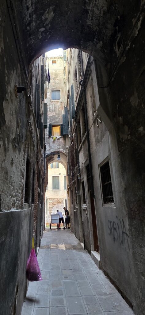 A very narrow Venetian alley running under a low arch, damp stained walls on both sides close enough to touch, washing hanging above, a purple bag on a hook and daylight opening onto a small square at the far end