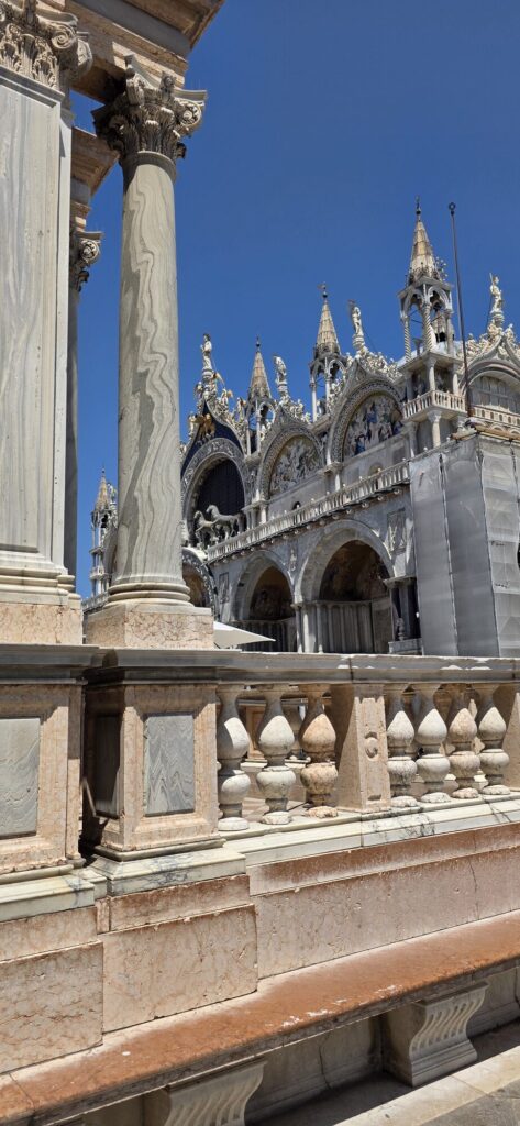 Saint Marks Basilica seen past the marble columns and stone balustrade of a loggia, its Gothic pinnacles and statues sharp against a deep blue sky