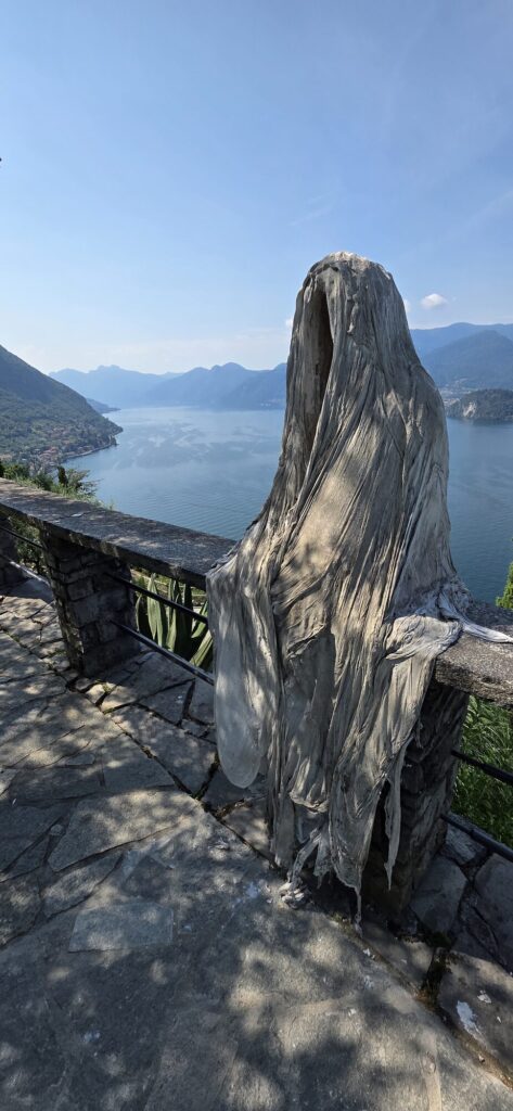 A life size hollow figure of stiffened white cloth standing on a stone terrace, hooded with an empty dark opening where a face would be, the folds falling in long vertical creases, with Lake Como and the mountains spread out behind
