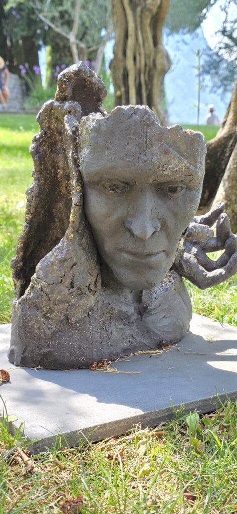 A fired clay sculpture of a face with a hand raised beside the ear, glazed grey with blackened and metallic patches and a crackled surface, standing on a slate base in the grass under an olive tree